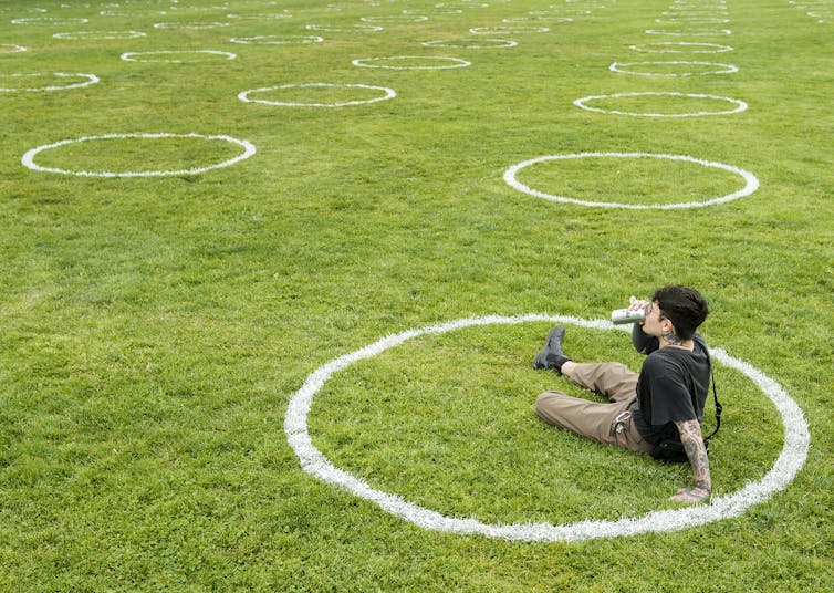 A man sits in a white circle painted on the grass at a park and drinks a beer