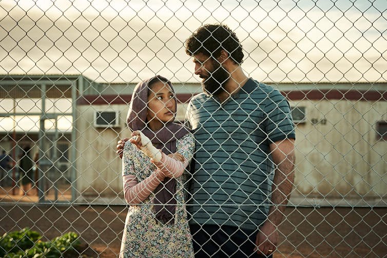 A Film Still, Set In An Immigration Detention Centre, A Woman And A Man Stand Behind A Fence.