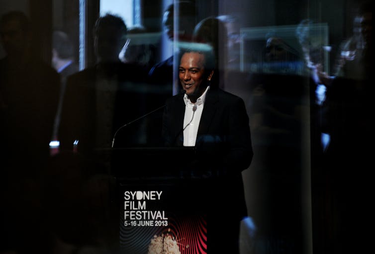 A man wears a suit and stands behind a lectern reading 'Sydney Film Festival'
