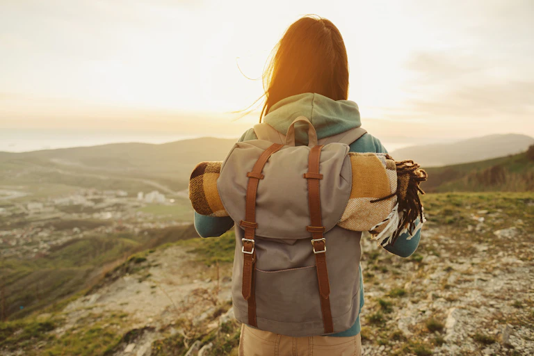 A woman wears a backpack, standing on top of a hill.