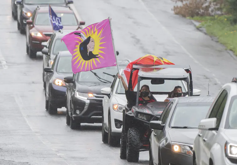 A convoy of vehicles with First Nations flags drive along a road