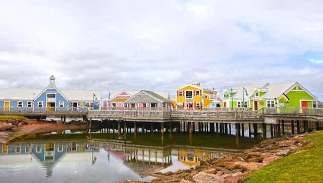 A blue, pink, yellow and green houses on a wooden pier over a small bay. The bay is lined with red rocks on top of which sit a green lawn. The sky behind the houses is filled with white clouds. 