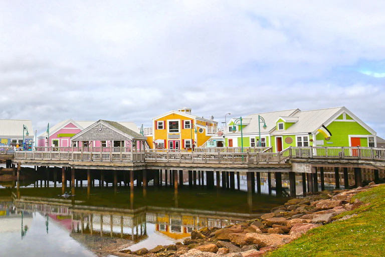 A blue, pink, yellow and green houses on a wooden pier over a small bay. The bay is lined with red rocks on top of which sit a green lawn. The sky behind the houses is filled with white clouds.