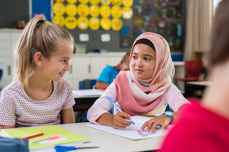 Two female students, one of whom wears a hijab, sit in a classroom talking.