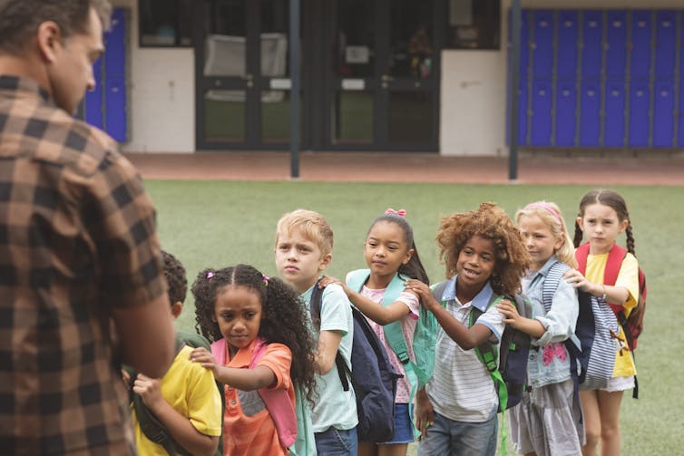 A teacher stands outdoors and faces a line of schoolchildren of diverse racialized backgrounds.