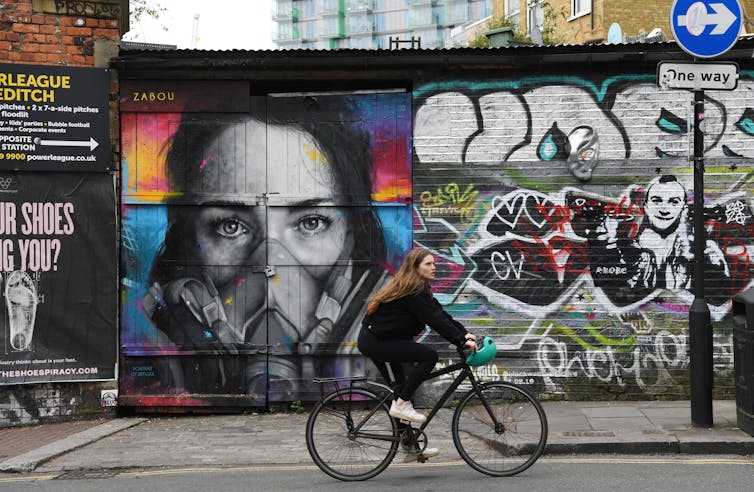 Woman cycles past street art of person wearing respirator.