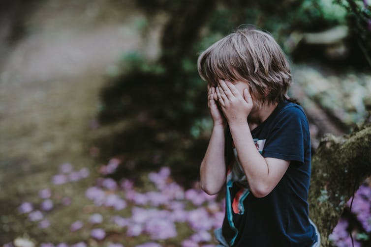 A boy weeps into his hands in an outdoor setting, with trees in the background.