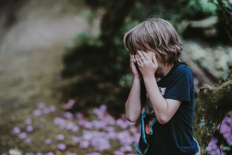 A boy weeps into his hands in an outdoor setting, with trees in the background.