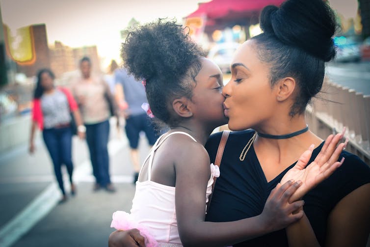 A mother kisses her young daughter in a public setting, with people in the background walking behind them.