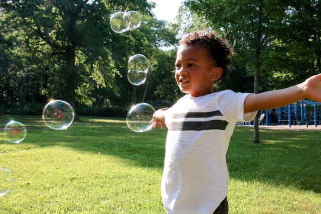A Black child stands in grass with a playground behind him with his arms open as bubbles float around him. 