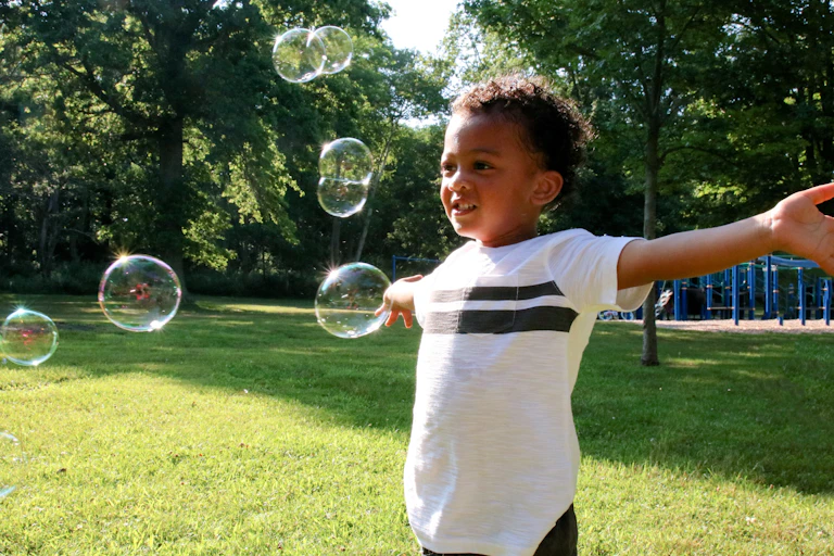A Black child stands in grass with a playground behind him with his arms open as bubbles float around him.