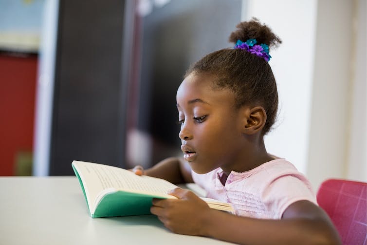A girl who is Black is reading a book at a school desk.