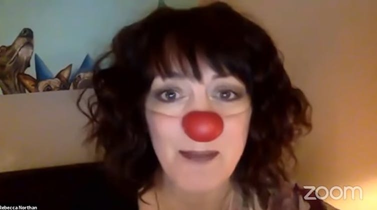 A woman in costume as a clown with a red nose looks with anticipation at viewers on a Zoom screen.