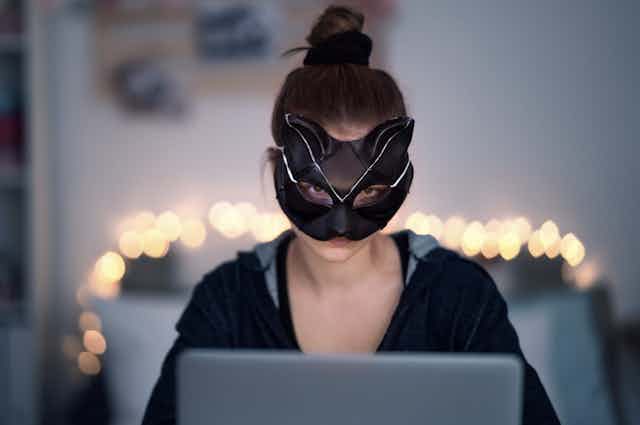 A woman wearing a cat mask sits in front of a laptop at night.