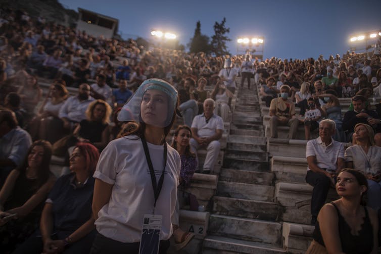 An usherette wears a face shield as she stands in front of rows of audience members in twilight at the Odeon of Herodes Atticus in Athens, Greece.