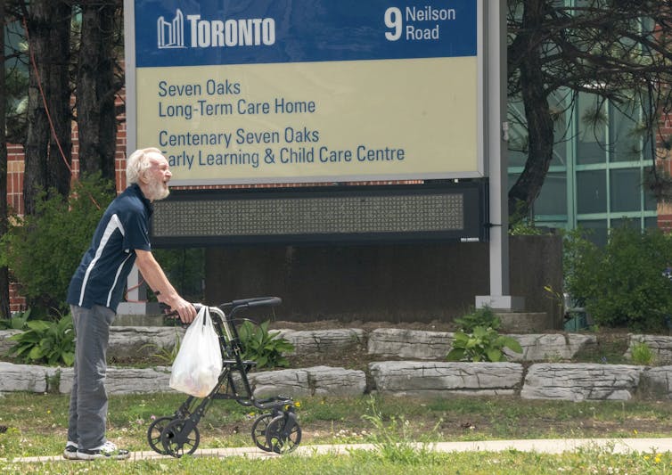 A smiling, bearded man toting a plastic bag and using a walker takes a walk outside a long-term care home in Toronto, with a sign with the name of the home — Seven Oaks — in the background.
