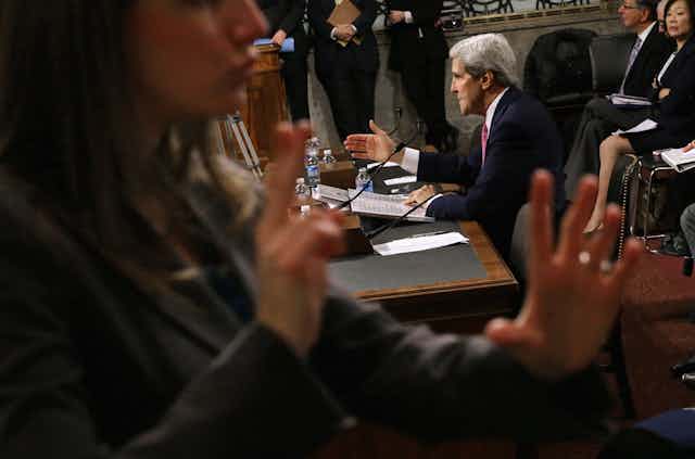 A woman signs as a seated John Kerry speaks to a crowded congressional chamber.