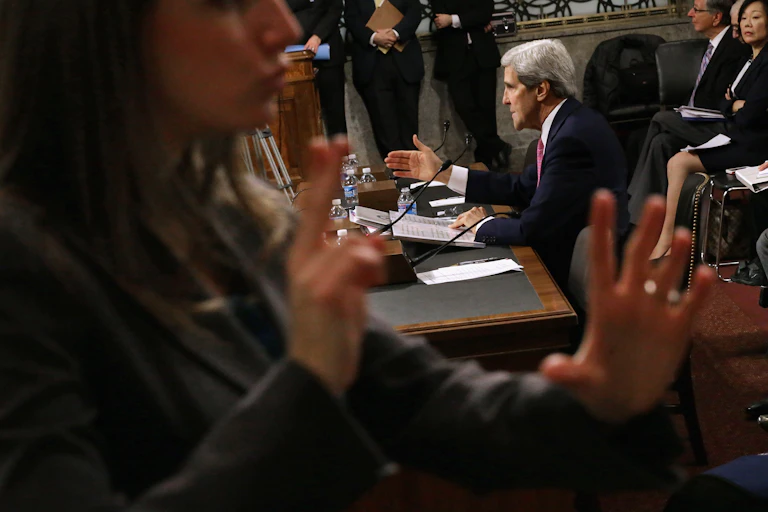 A woman signs as a seated John Kerry speaks to a crowded congressional chamber.