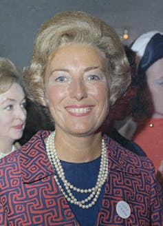 Vera Lynn stands smiling wearing a triple string of pearls and a jacquard blue and orange jacket in 1969.