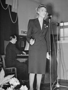 Vera Lynn stands at a microphone singing, backed by a pianist.