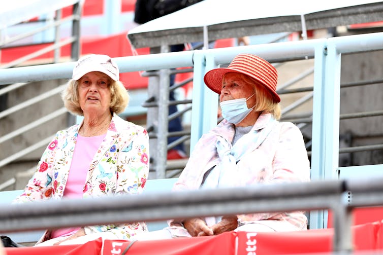 Two older women in spectator seats, one without mask, one worn improperly