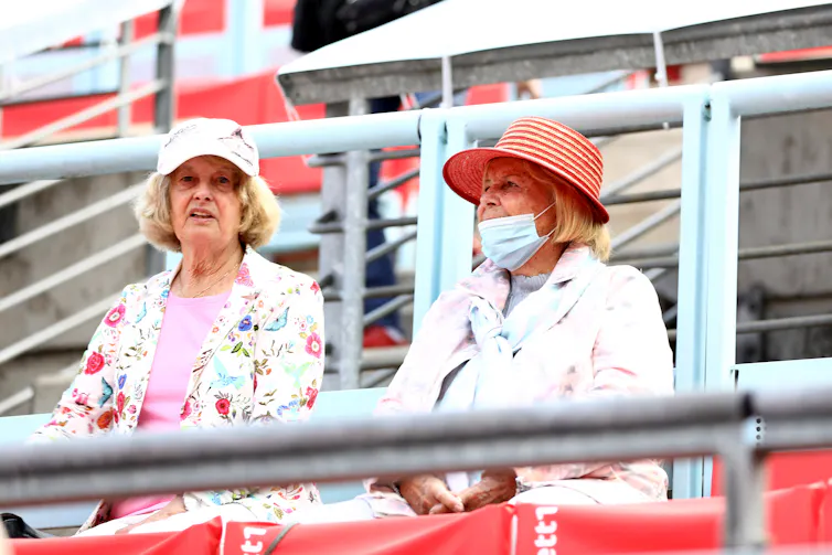 Two older women in spectator seats, one without mask, one worn improperly