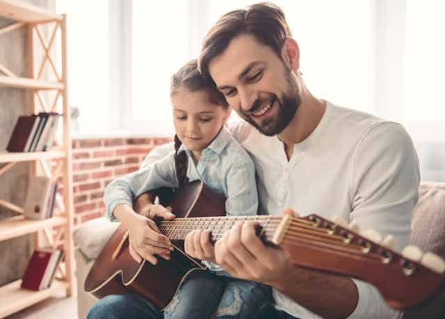 A father and his daughter hold a guitar together in a living room. There is a wooden shelf in front of a low brick wall.