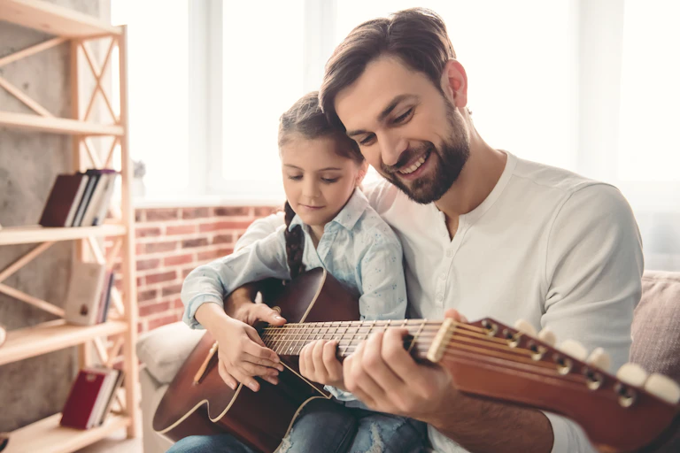 A father and his daughter hold a guitar together in a living room. There is a wooden shelf in front of a low brick wall.