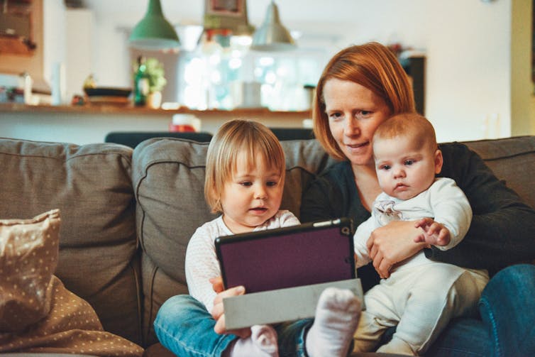 A mom holds two kids in her lap on a brown couch as they look at a tablet.