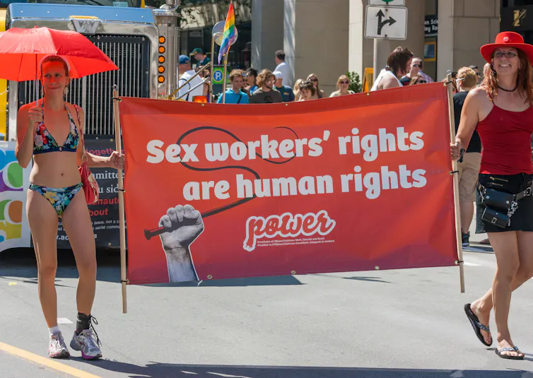 Two women, one in a bikini carrying a red umbrella, another in a red tank top, black skirt and red cowboy hat, carry a banner between them that says, 'Sex workers' rights are human rights.' There is a crowd a few meters behind them.
