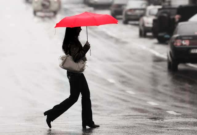 A woman walks across a busy road holding a red umbrella.