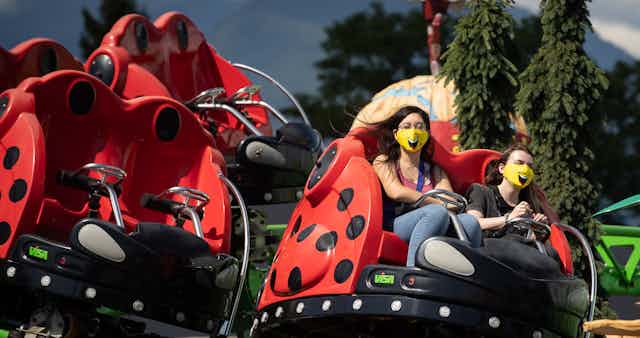 Two women ride a spinning amusement park ride while wearing yellow smiley-face face masks with evergreen trees and mountains in the background.