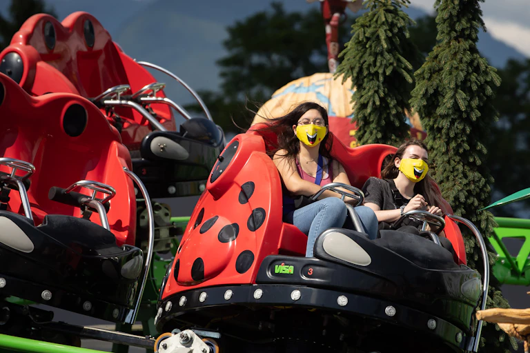 Two women ride a spinning amusement park ride while wearing yellow smiley-face face masks with evergreen trees and mountains in the background.