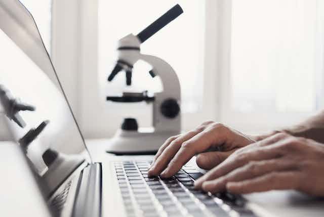 In the foreground, two hands rest on a laptop keyboard. In the background, a slightly out-of-focus microscope.