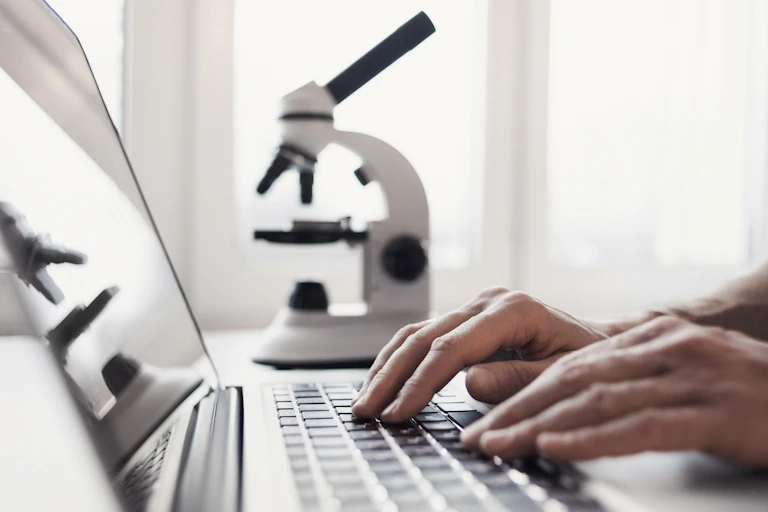 In the foreground, two hands rest on a laptop keyboard. In the background, a slightly out-of-focus microscope.