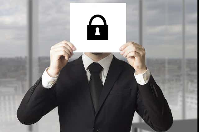 A man in a black business suit holds up a white card with a black symbol of a padlock, obscuring his face.