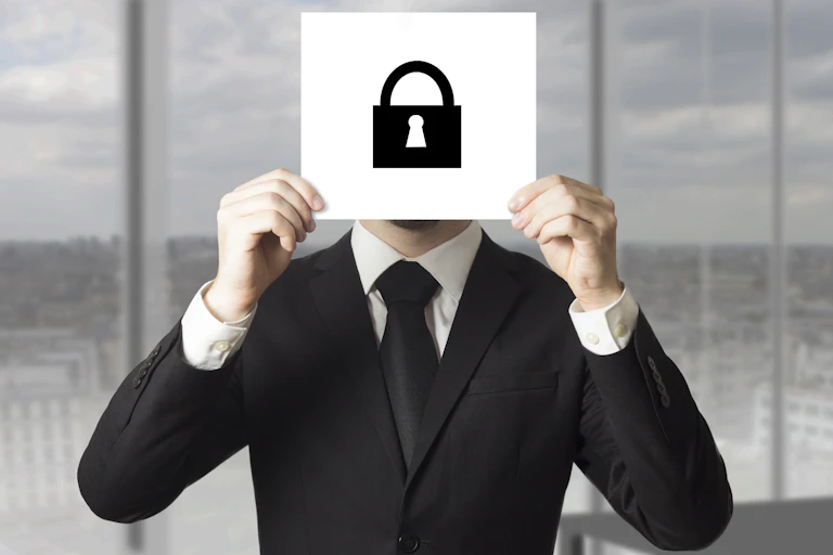 A man in a black business suit holds up a white card with a black symbol of a padlock, obscuring his face.