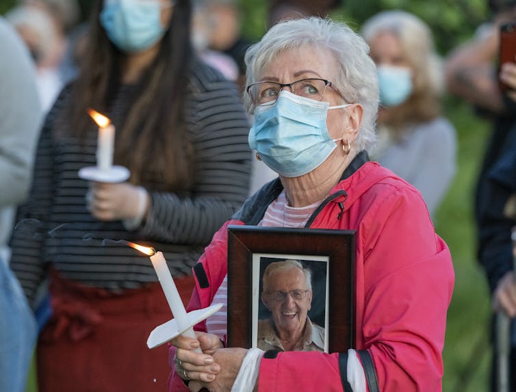 A grey-haired woman wears a mask and carries a candle and a framed photo of an elderly man at a vigil.