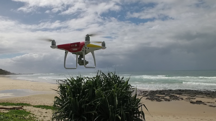 A red and white drone hovers by an empty beach.