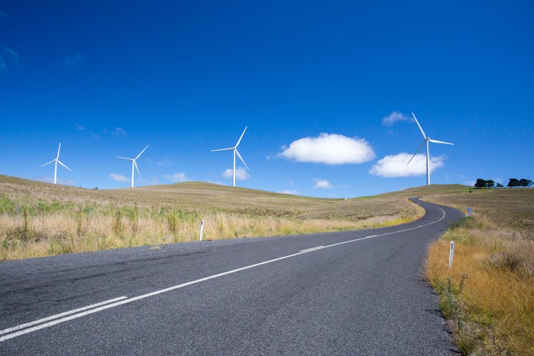 Four wind turbines at the end of a road, flanked by pale green-brown grass.