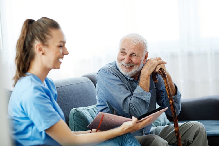 Elderly man sits on the couch happily talking with young female carer.