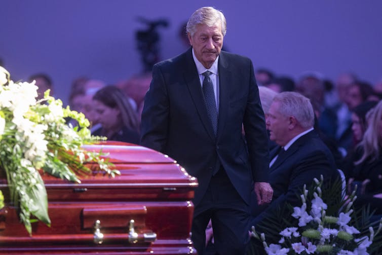 Mike Harris, in suit, tie and overcoat, is seen walking past a casket covered with white flowers.