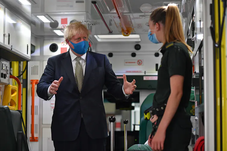 Prime Minister Boris Johnson, wearing a face mask, talks with a paramedic as they stand inside the back of an ambulance.