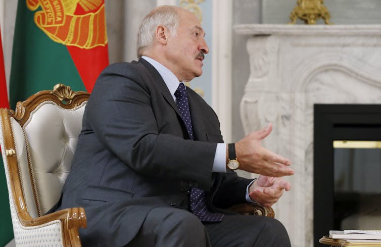 Alexander Lukashenko sitting on a chair with flag behind.