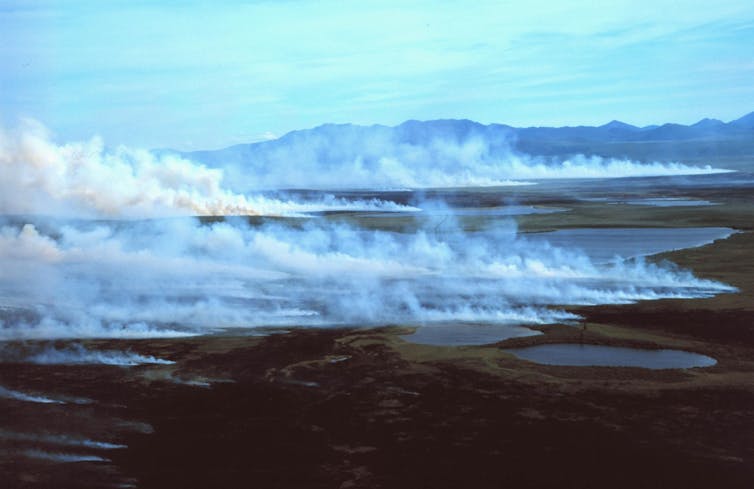 White smoke rises from the tundra with mountains in the background.