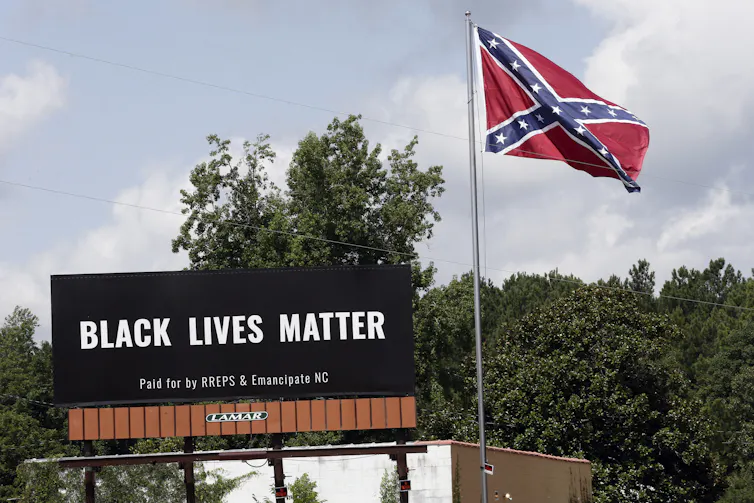 A Black Lives Matter billboard sits next to a Confederate flag, with trees in the background.