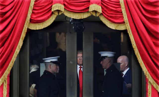 Trump peers through a space between two doors, flanked by military, as he waits to make his inaugural address. Red bunting with gold tassels envelop the scene.
