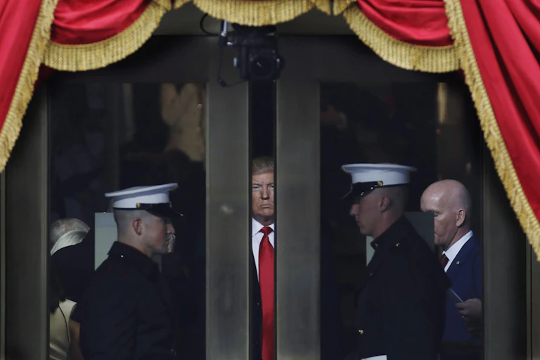 Trump peers through a space between two doors, flanked by military, as he waits to make his inaugural address. Red bunting with gold tassels envelop the scene.
