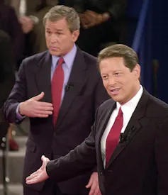 Al Gore is seen gesturing in a debate with George W. Bush, as Bush looks on with a 'who me?' expression in the background.