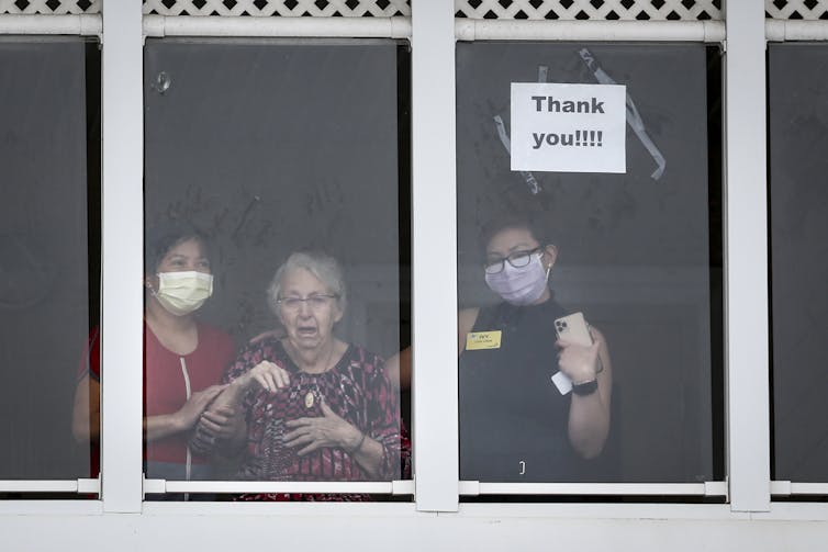 An elderly woman, flanked by two long-term health-care workers wearing masks, looks out a window and waves.